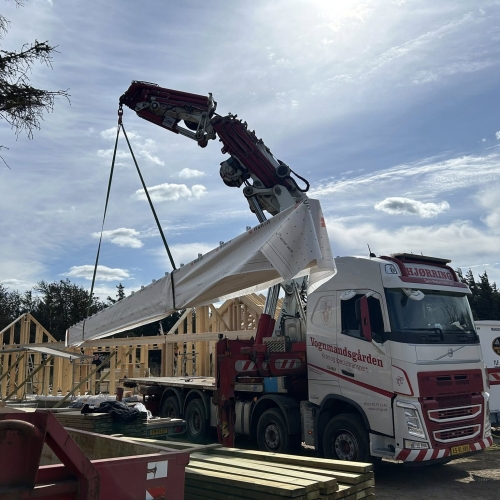 Krankørsel med stor træbjælke løftet over lastbil ved nybyggeri.