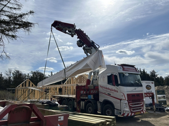 Krankørsel med stor træbjælke løftet over lastbil ved nybyggeri.